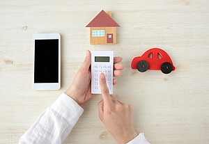 There is a smartphone, a small house and a small car on a light table. A man calculates on a calculator©Getty Images/takasuu via canva pro