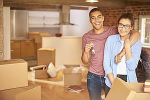  A young couple smiles into the camera. They hug each other. Behind them there are moving boxes in the empty apartment © iStock/sturti