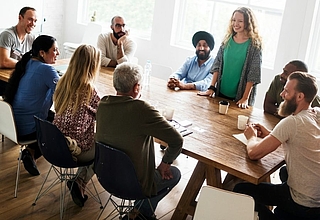  9 people sit at a wooden table and discuss ©Getty Images Pro_Rawpixel Ltd via Canva Pro
